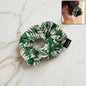 A green and white leaf print scrunchie with a black 'viosi' tag rests on a marble surface, with an inset showing it in a bun.