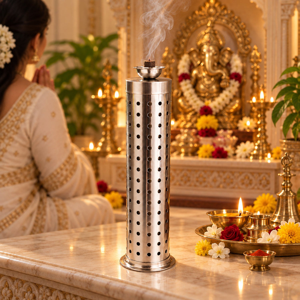 A stainless steel incense holder with smoke rising sits on a table adorned with lit lamps, flowers, and a golden deity