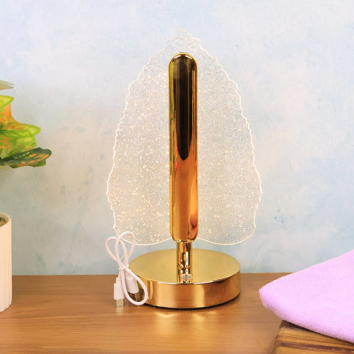 Crystal leaf lamp with a gold base and stem, glowing on a light wooden surface with a plant.