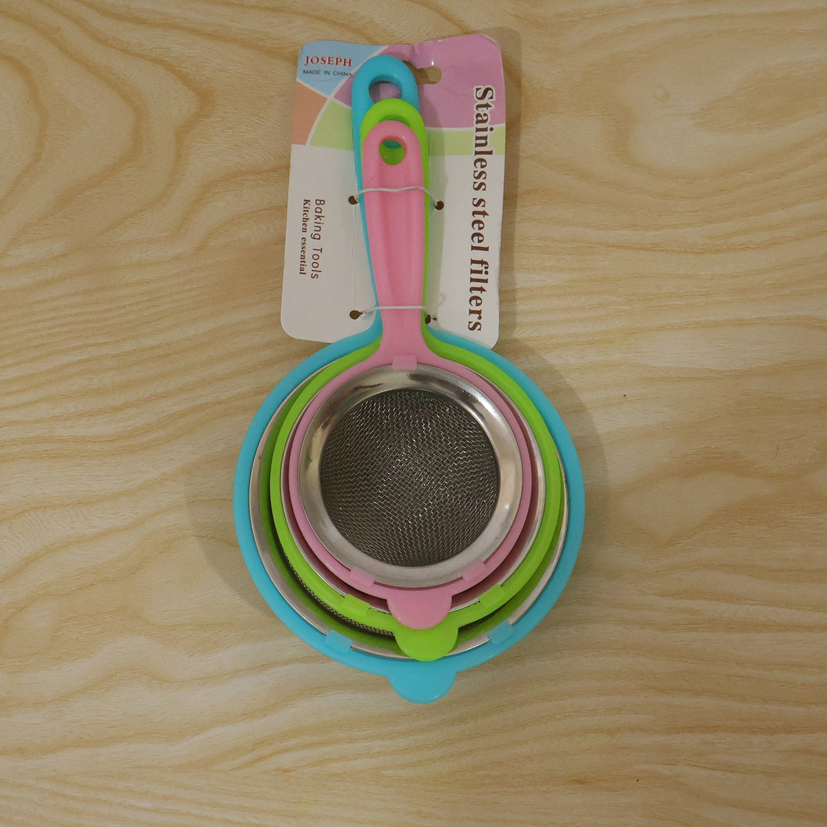 Three nested strainers with colorful plastic handles and stainless steel mesh are packaged on a wooden surface.