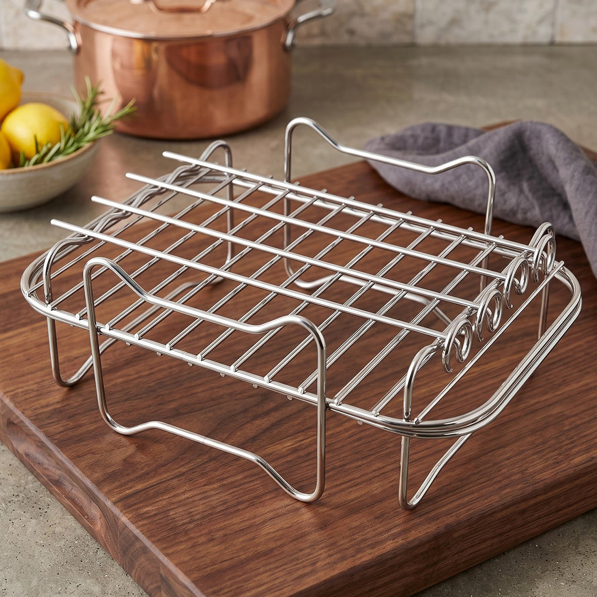 A shiny metal air fryer rack sits on a dark wooden cutting board, with a copper pot and lemons in the background.