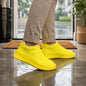 A person wearing khaki pants and bright yellow silicone shoe covers stands on a wet tiled floor near a doorway.