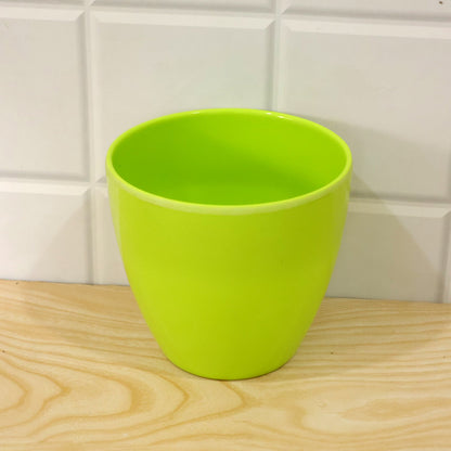 An empty, lime green plastic pot resting on a light wood surface, with white tiled wall in the background.