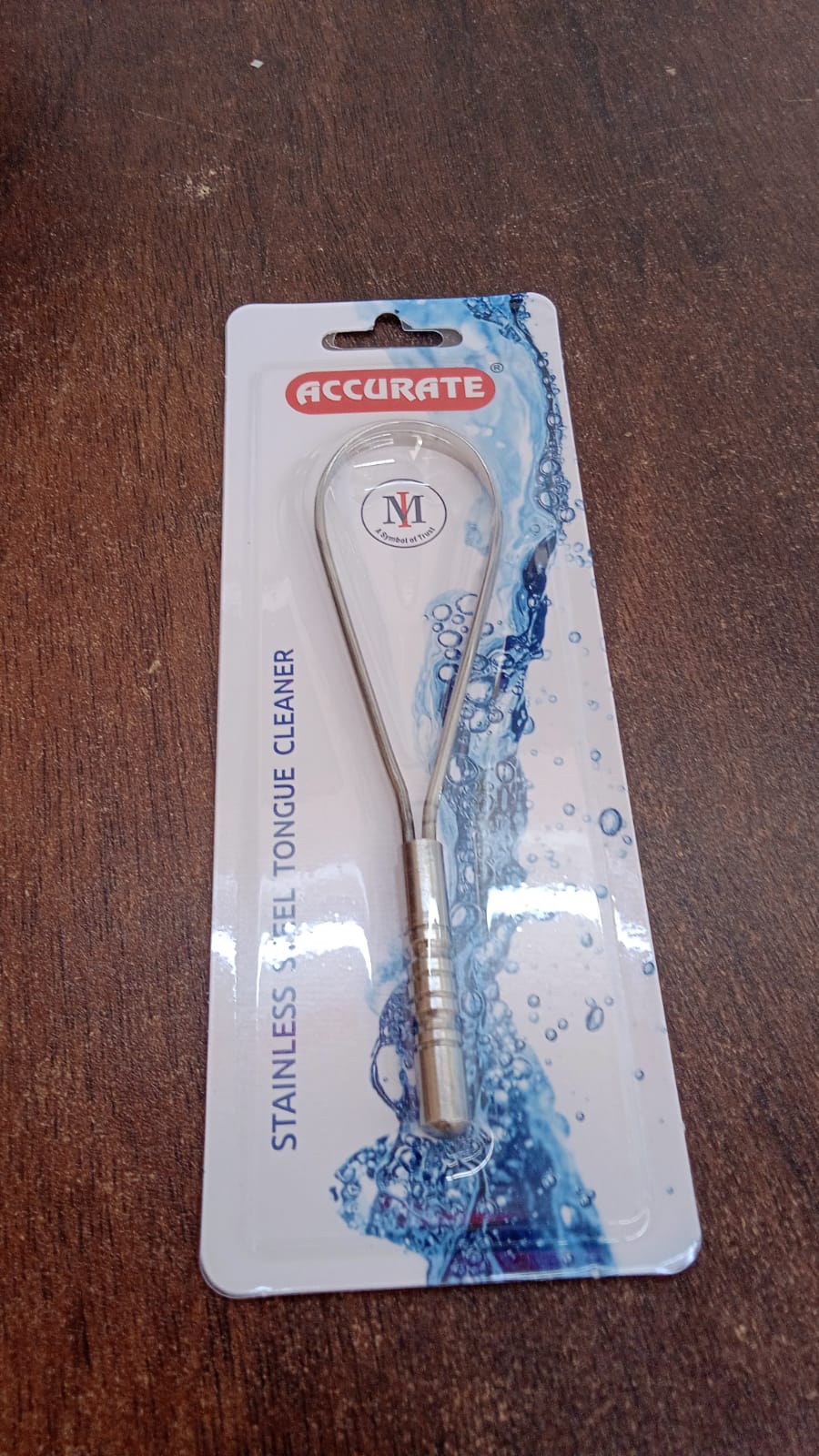 A stainless steel tongue cleaner in clear plastic packaging with a blue water design rests on a wooden surface.