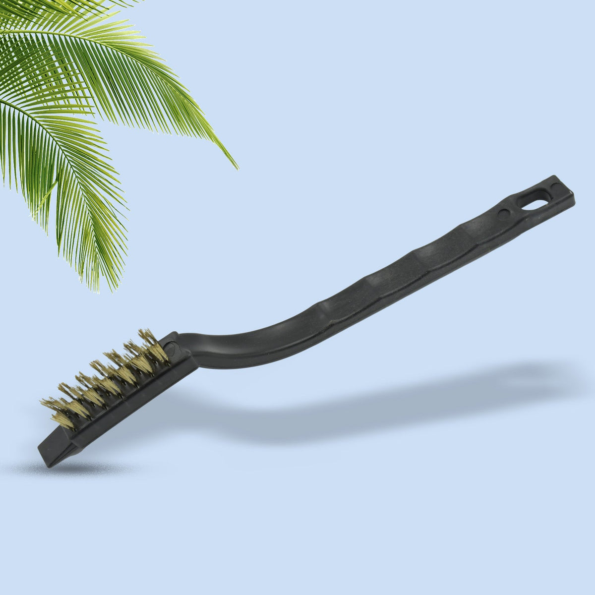 A single black-handled mini wire brush with brass bristles, casting a shadow on a light blue background with a palm leaf in