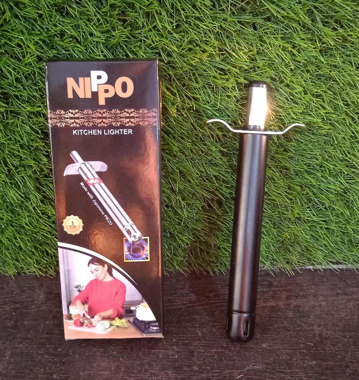 A black and silver gas lighter stands next to its black packaging, against a green grassy background.
