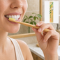 A woman brushes her teeth with a bamboo toothbrush, a packaged toothbrush in the background.