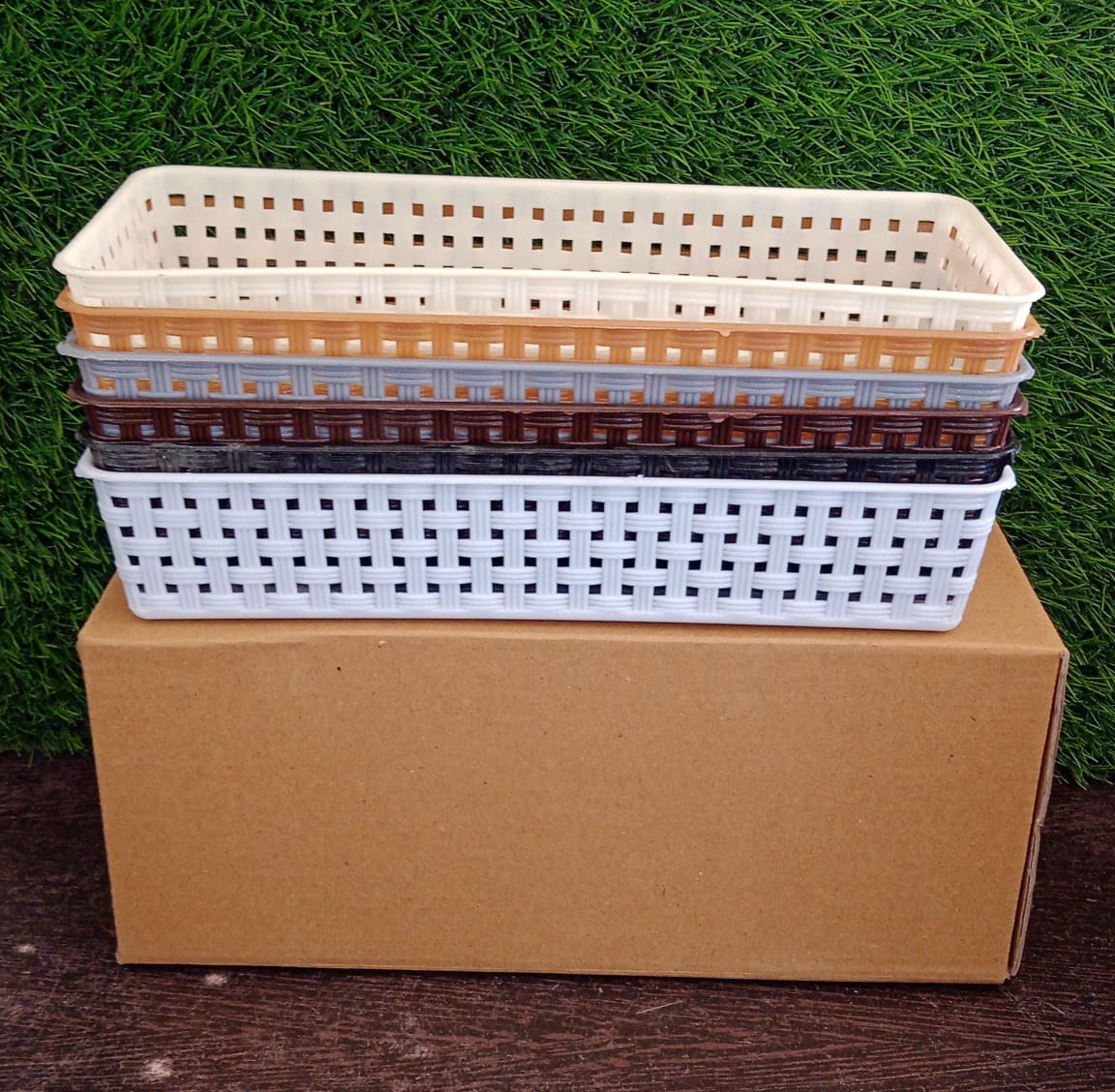 A stack of six rectangular plastic baskets in various colors on a brown cardboard box with a green background.
