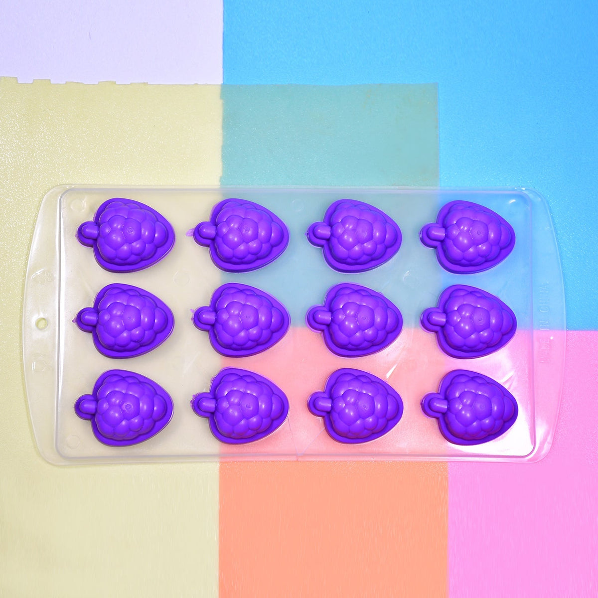 Purple silicone fruit-shaped ice cube tray on a colorful background of blue, pink, and yellow squares.