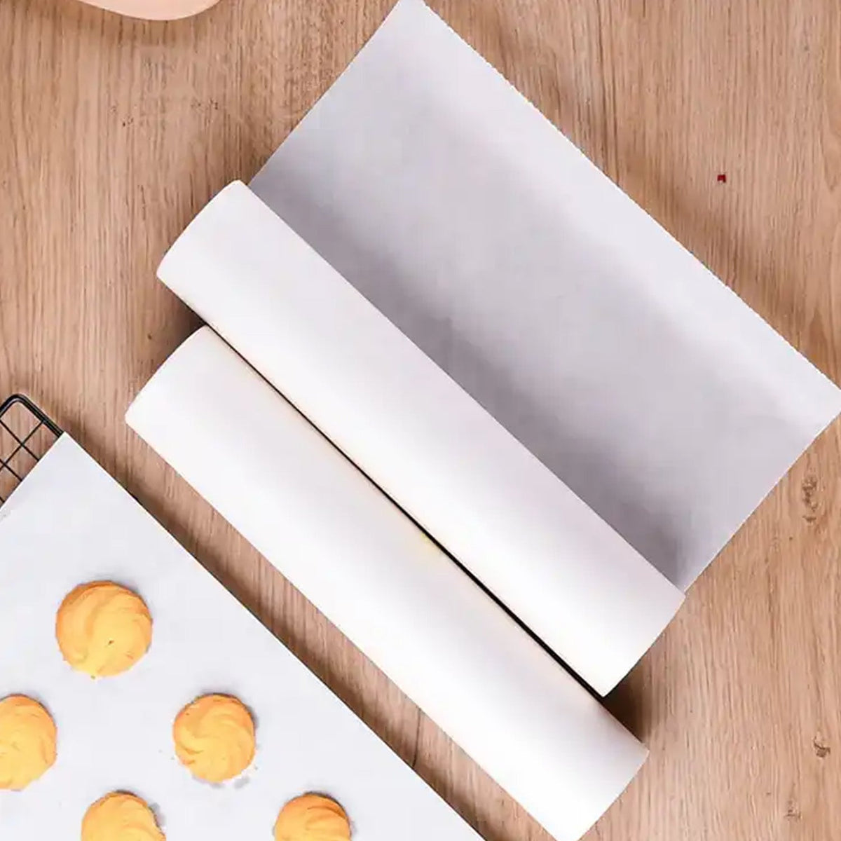 Two rolls of white non-stick baking paper on a wooden surface next to a baking sheet with cookies on a piece of the paper.