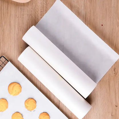 Two rolls of white non-stick baking paper on a wooden surface next to a baking sheet with cookies on a piece of the paper.