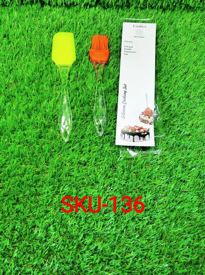 A yellow silicone spatula, an orange silicone brush, and a packaged silicone cooking set lie on artificial green grass.