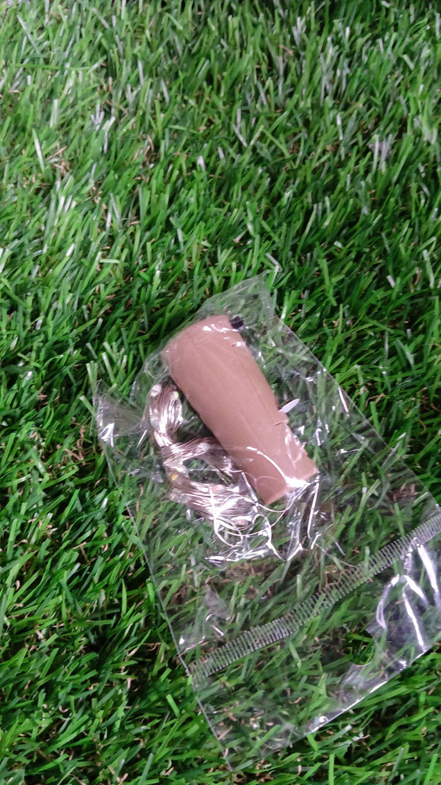 A single bottle cork light with coiled LED string lights, packaged in a clear plastic bag, resting on artificial green grass.
