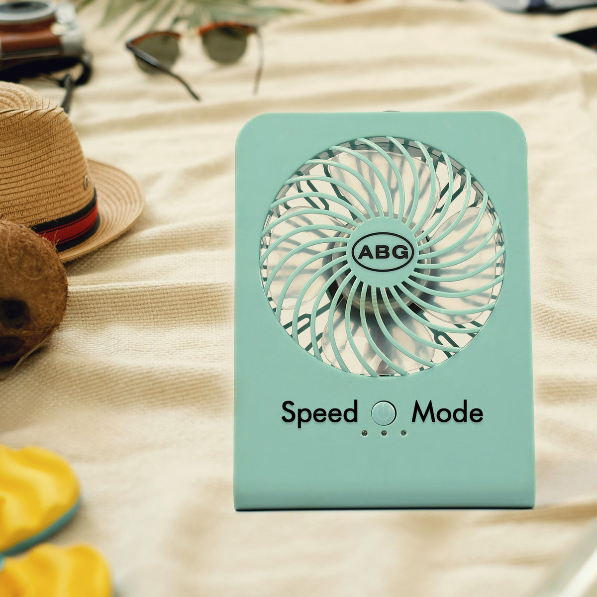 A mint green portable desk fan on a beige blanket, surrounded by a straw hat, sunglasses, and coconut halves.