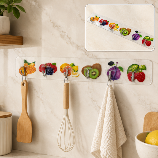 A white adhesive wall hook strip with fruit designs holds a wooden spatula, whisk, and white towel in a kitchen.