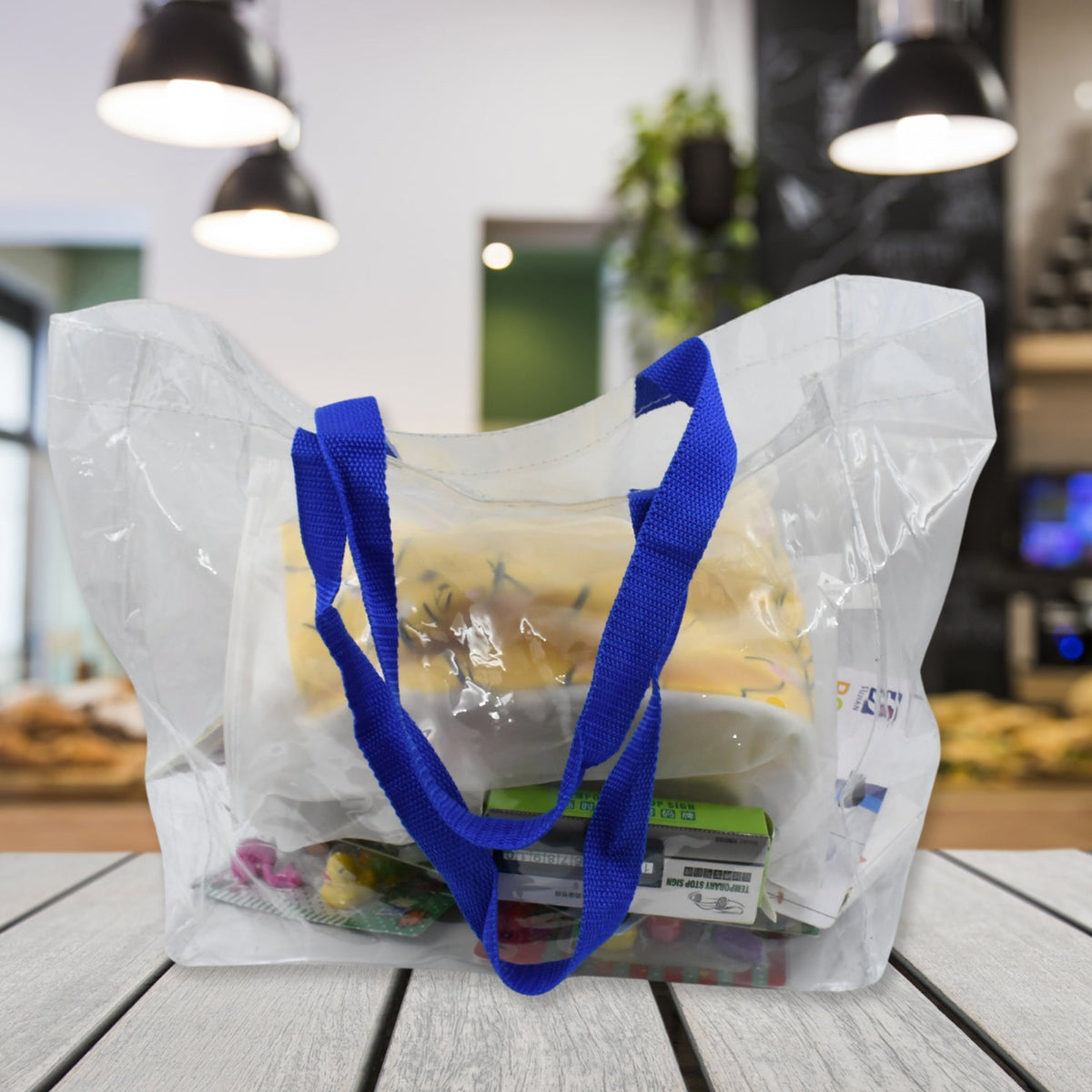 A clear PVC tote bag with blue handles is filled with various items, sitting on a wooden counter in a blurred cafe.