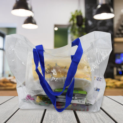 A clear PVC tote bag with blue handles is filled with various items, sitting on a wooden counter in a blurred cafe.