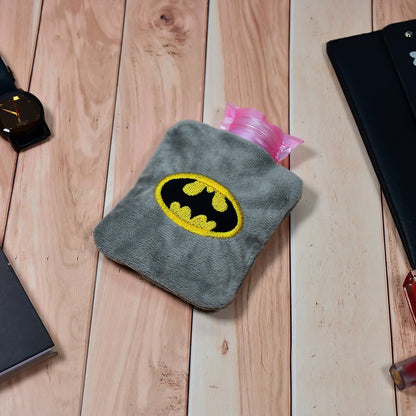 Gray plush hot water bag with a Batman logo on a wooden table with a watch and notebook.