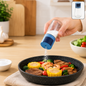 A hand holds a blue and clear glass spice dispenser, shaking salt over a pan of cooked vegetables and meat.