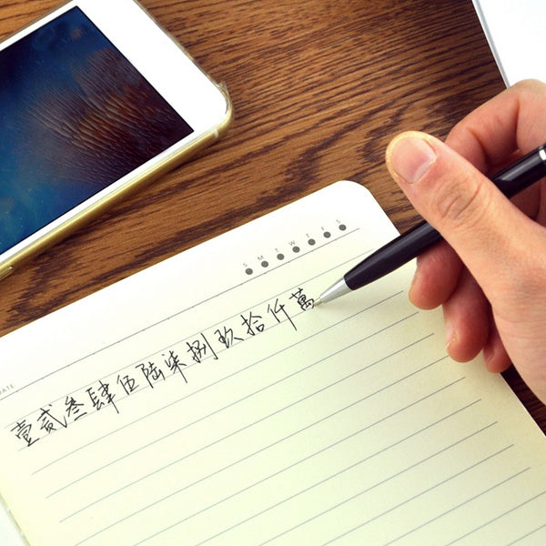 A hand holding a black 3-in-1 stylus pen, writing in a notebook next to a smartphone.