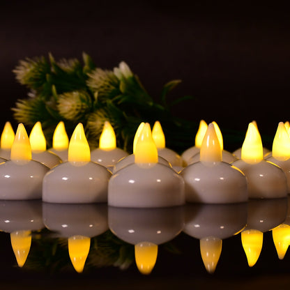 A close-up of multiple white LED floating candles with yellow flames, reflected on a dark surface, with green foliage behind.