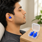 A man with closed eyes wears blue silicone earplugs while resting, with a clear case of earplugs on a table.