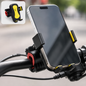 Black and yellow phone mount attached to a bicycle handlebar, holding a silver smartphone. Inset shows the mount without