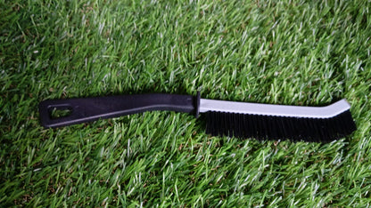 A black and gray crevice cleaning brush with black bristles rests horizontally on a vibrant green artificial grass surface.