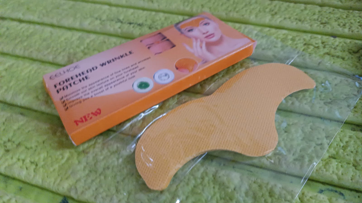 An orange box of forehead wrinkle patches next to a single gold patch in clear packaging on a green textured surface.