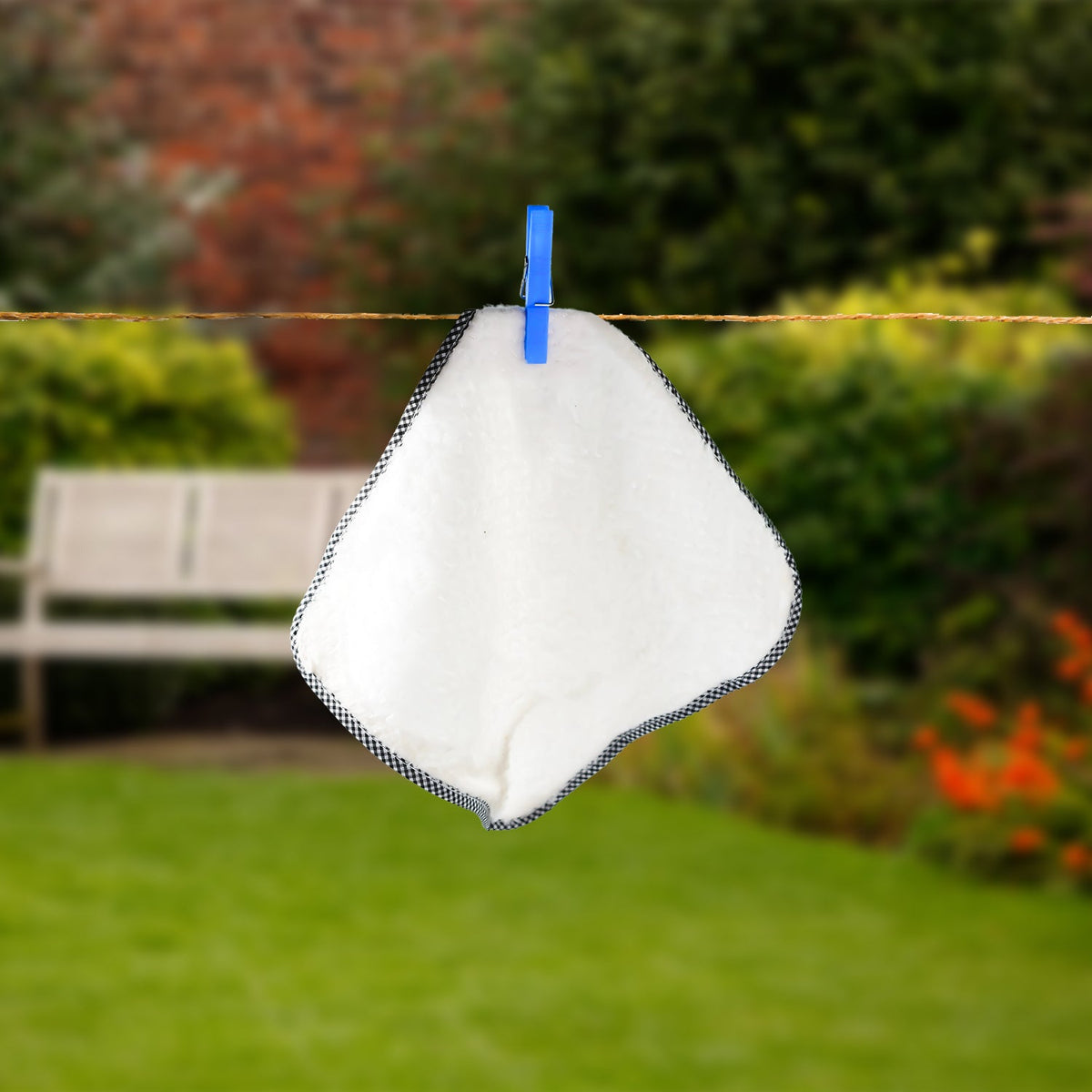 A white washcloth with a black and white trim hangs from a blue plastic clothes clip on a clothesline in a garden.