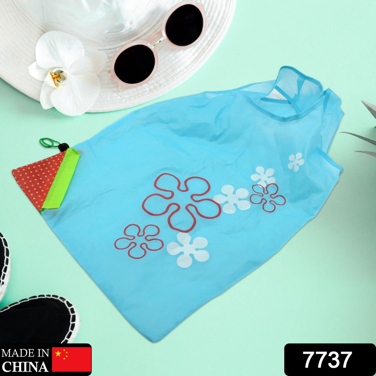 A light blue reusable shopping bag with red and white flower designs lies on a green surface next to sunglasses and a hat.