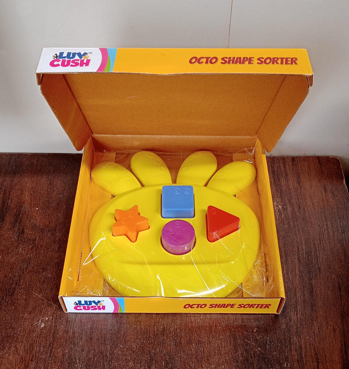 Yellow shape sorter toy with colorful blocks packaged inside an open cardboard box on a dark wooden surface.