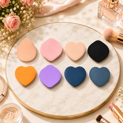 Eight colorful mini makeup sponges in various shapes on a marble tray with cosmetics around them.