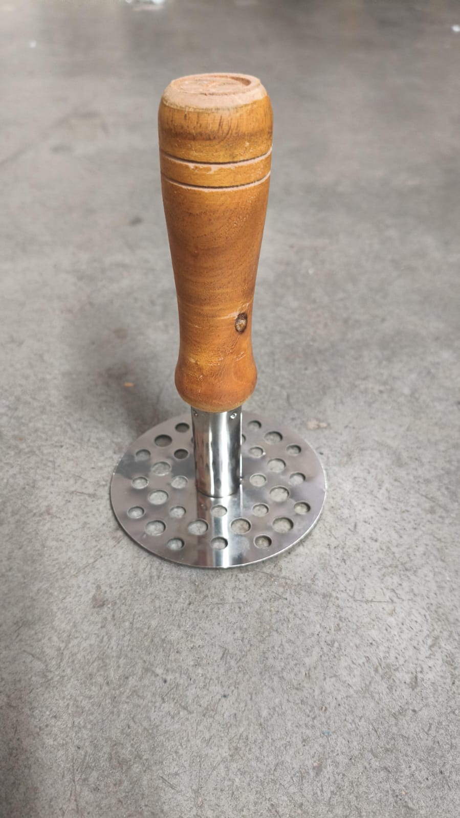 A stainless steel masher with a brown wooden handle rests on a gray concrete surface.