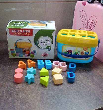 A blue and yellow shape sorter bucket and colorful blocks are displayed next to their product box, which reads "BABY'S FIRST