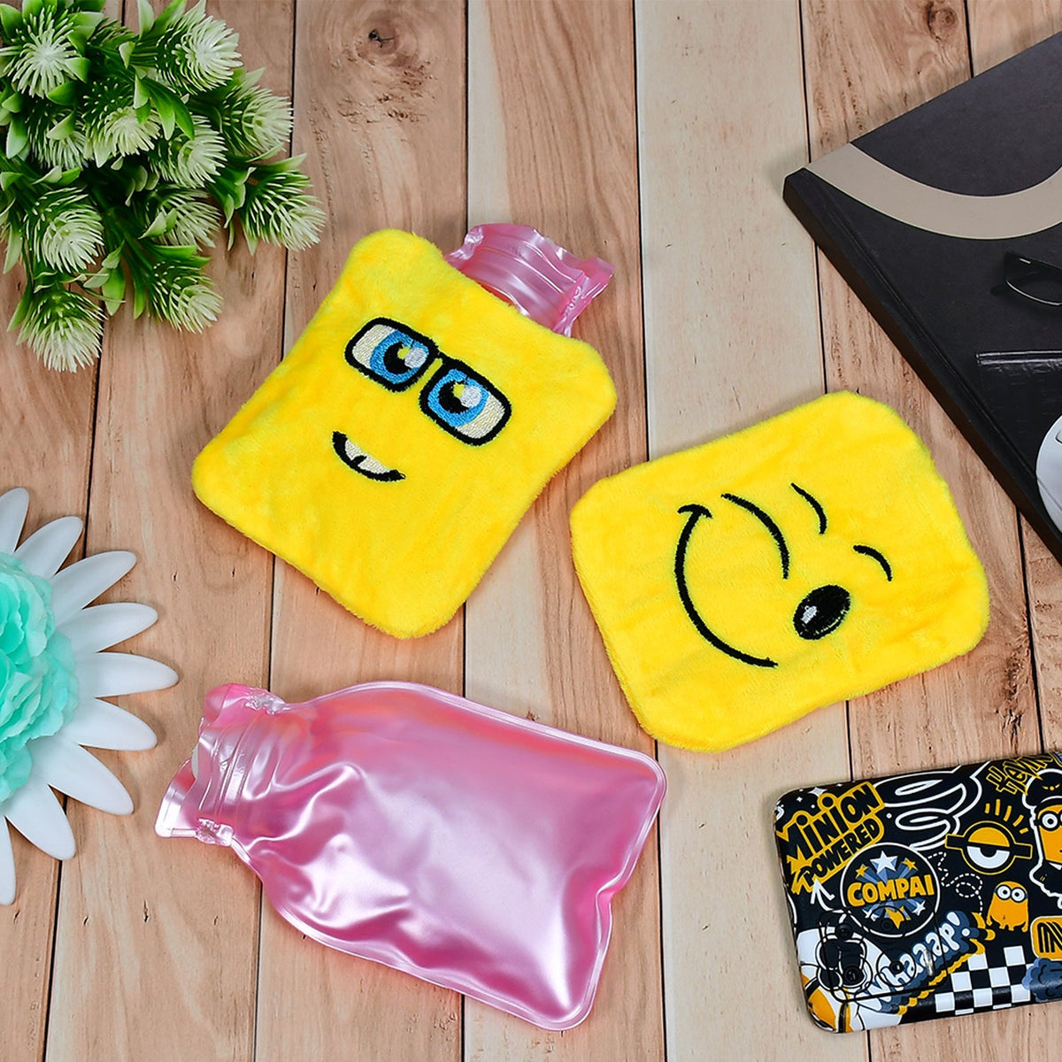 Two yellow emoji hot water bags and one pink plastic hot water bottle are on light wooden surface.