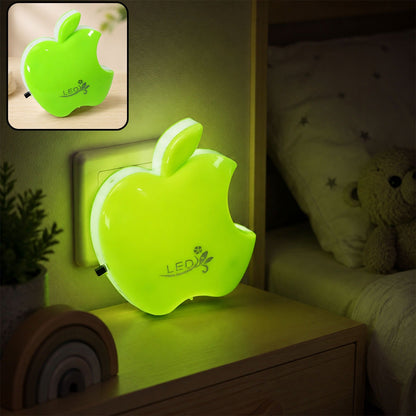 Green apple-shaped LED night light plugged into a wall socket, illuminating a bedside table.