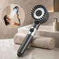 A dark gray shower head with a wide face and rubber bristles, next to a rolled towel, with an inset of a woman showering.