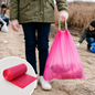 A person wearing gloves holds a full pink drawstring garbage bag on a sandy beach.