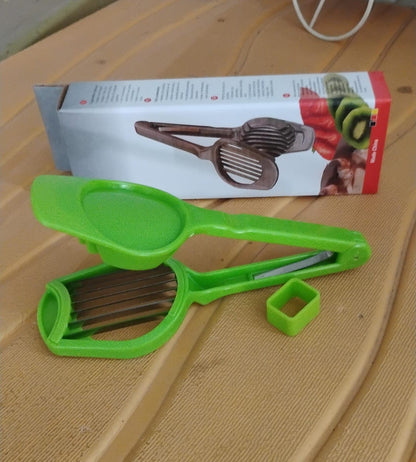 A green plastic slicer with metal blades and a small green square tool rests on a textured surface next to its product box.