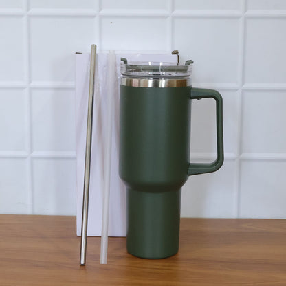 A dark green insulated travel mug with a handle, clear lid, and two straws stands on a wooden surface in front of a white