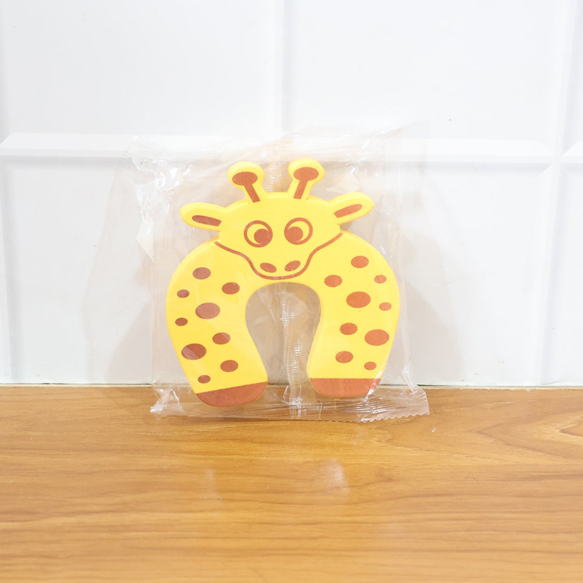 A yellow giraffe-shaped EVA door stopper is sealed in clear plastic packaging, resting on a wooden floor.