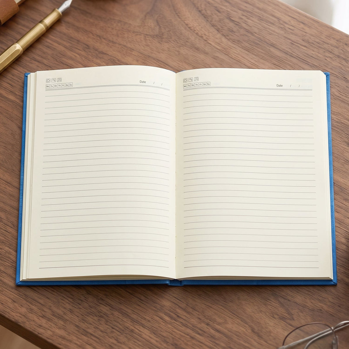 An open blue hardbound ruled diary with blank lined pages and date fields at the top, on a wooden desk with a pen nearby.