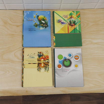 Four different hardbound ruled notebooks with colorful covers featuring various designs, arranged on a light wooden surface.
