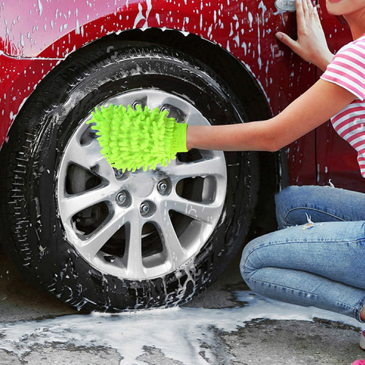 A person in jeans and a striped shirt washes a red car's wheel with a bright green microfiber glove.