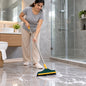 A woman in a grey shirt and beige pants scrubs a tiled bathroom floor with a green and yellow brush, creating white foam.