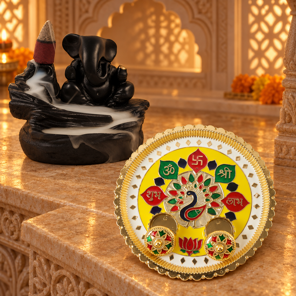 A black Ganesha backflow incense burner with smoke and a yellow and gold puja thali with peacock design on a marble surface.