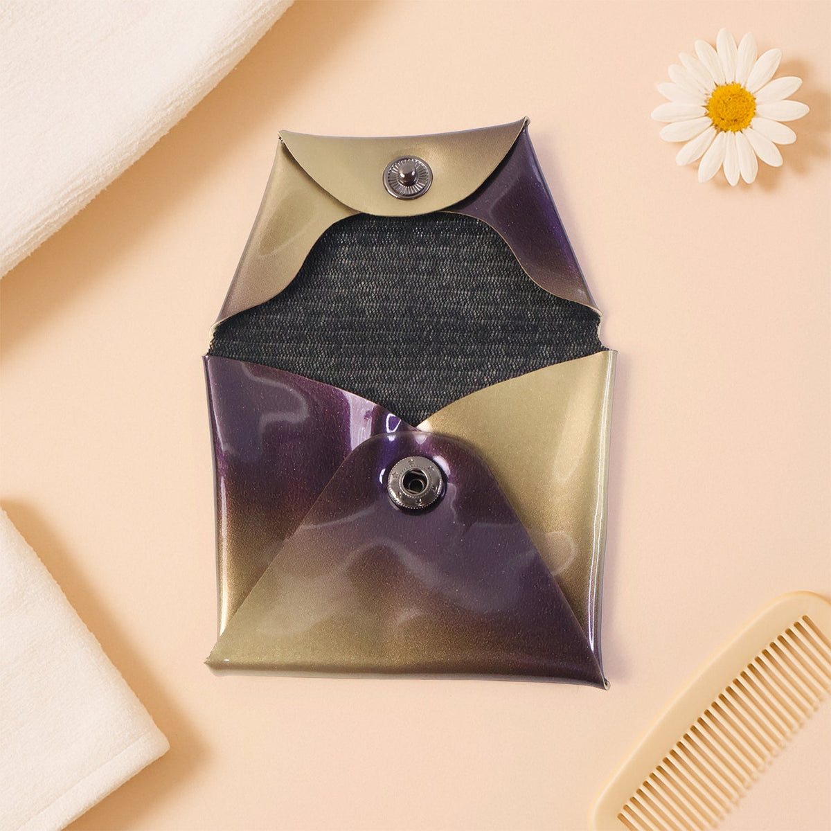 An open iridescent coin pouch with purple and gold hues lies on a peach surface next to a comb and towel.