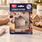 Three stainless steel house-shaped cookie cutters in clear packaging, surrounded by baked gingerbread cookies and baking