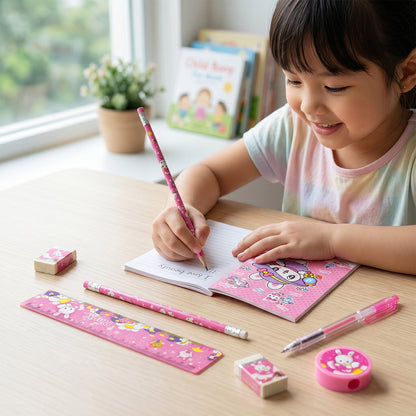 A smiling young girl writing in a small notebook with a pink pencil, surrounded by the matching stationery set on a wooden